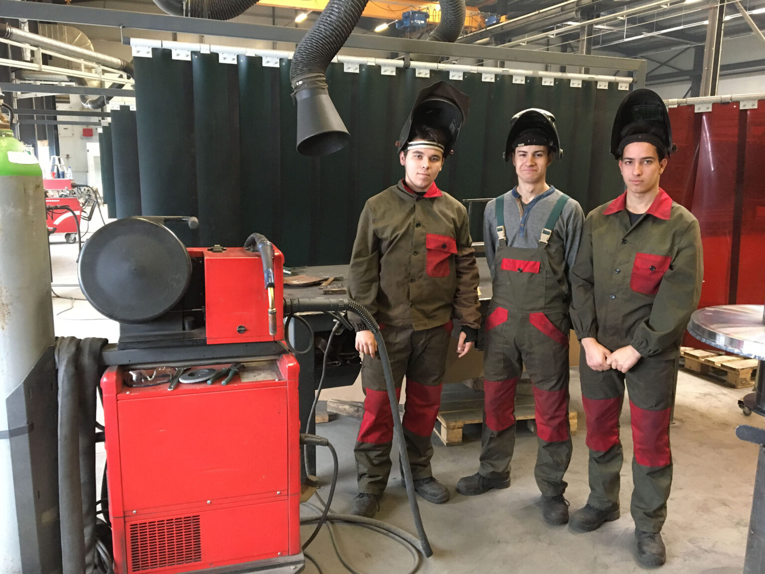 Três trabalhadores com equipamento de proteção estão ao lado de uma máquina de soldadura numa oficina.