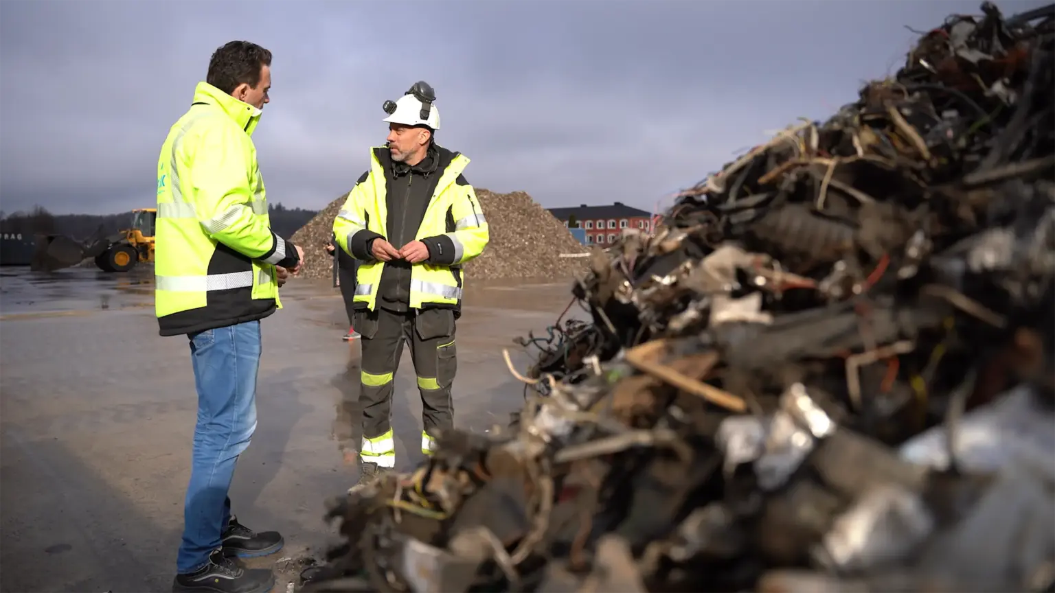 Dois homens com equipamento de proteção estão a discutir em frente a uma pilha de sucata na Skrotfrag, uma empresa de reciclagem de metais não ferrosos.