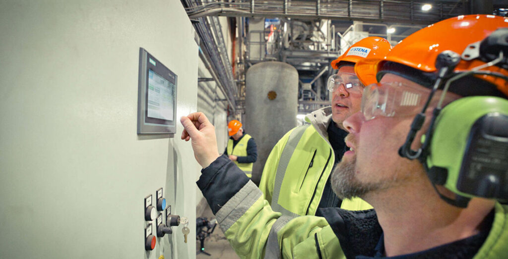 Dois trabalhadores com equipamento de proteção estão a operar um painel de controlo numa instalação industrial.