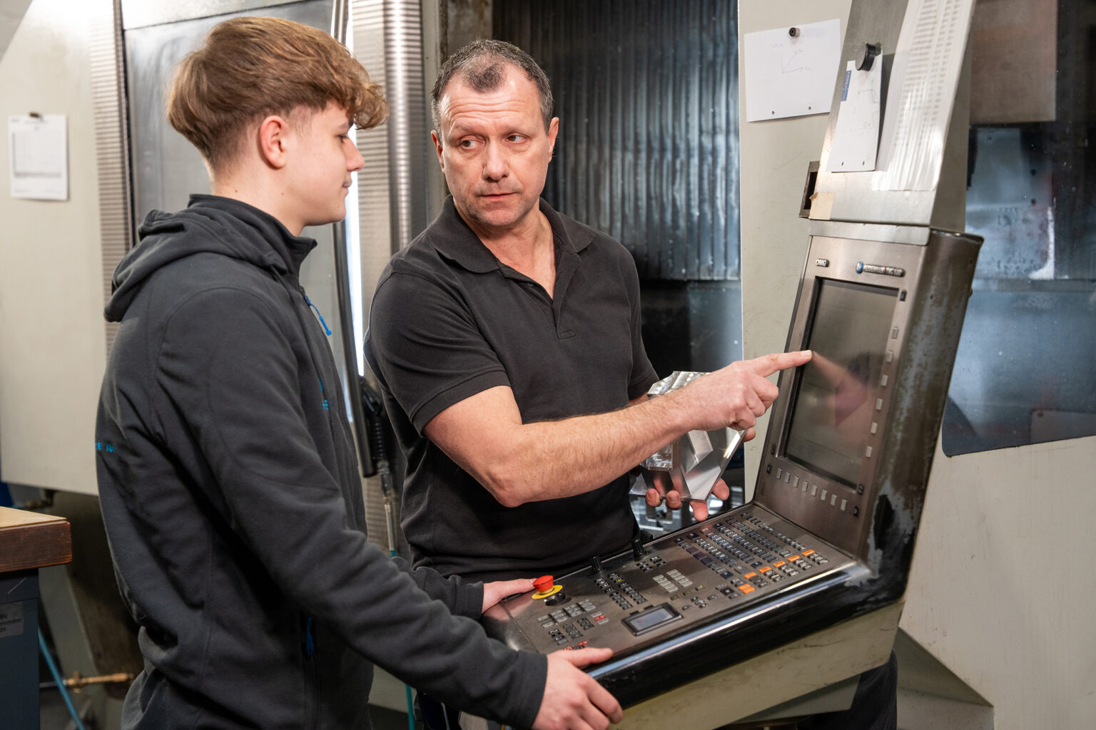 Um formador explica o funcionamento de uma máquina CNC a um jovem durante a formação para técnico de usinagem.