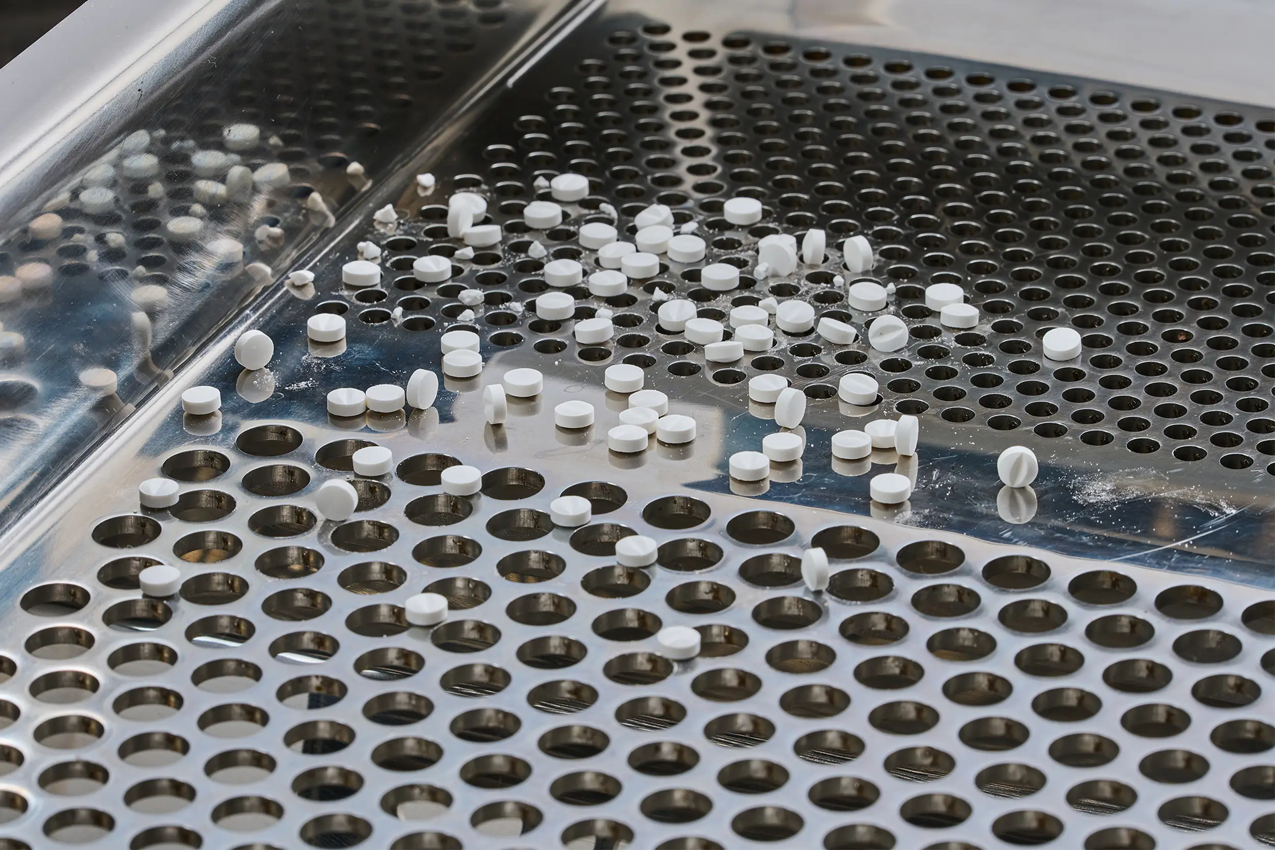 Comprimidos brancos numa peneira metálica, dentro de uma peneiradora farmacêutica.