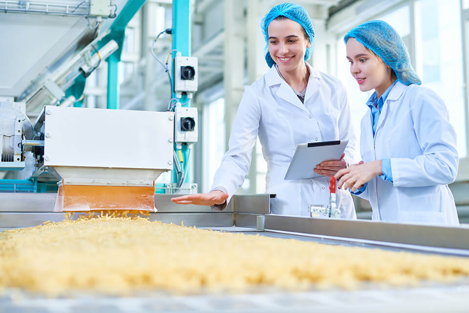 Duas mulheres vestidas com batas de laboratório inspecionam uma linha de produção com material a granel.