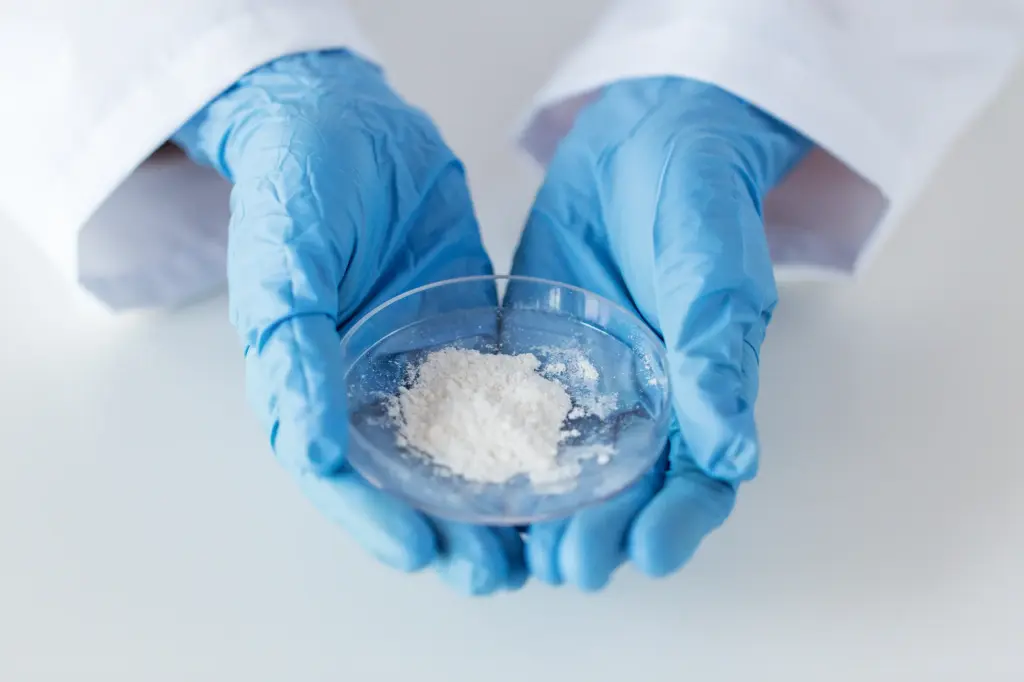 Imagem detalhada de uma placa de Petri com um pó branco, segurada por duas mãos com luvas de proteção — como imagem simbólica para produtos químicos