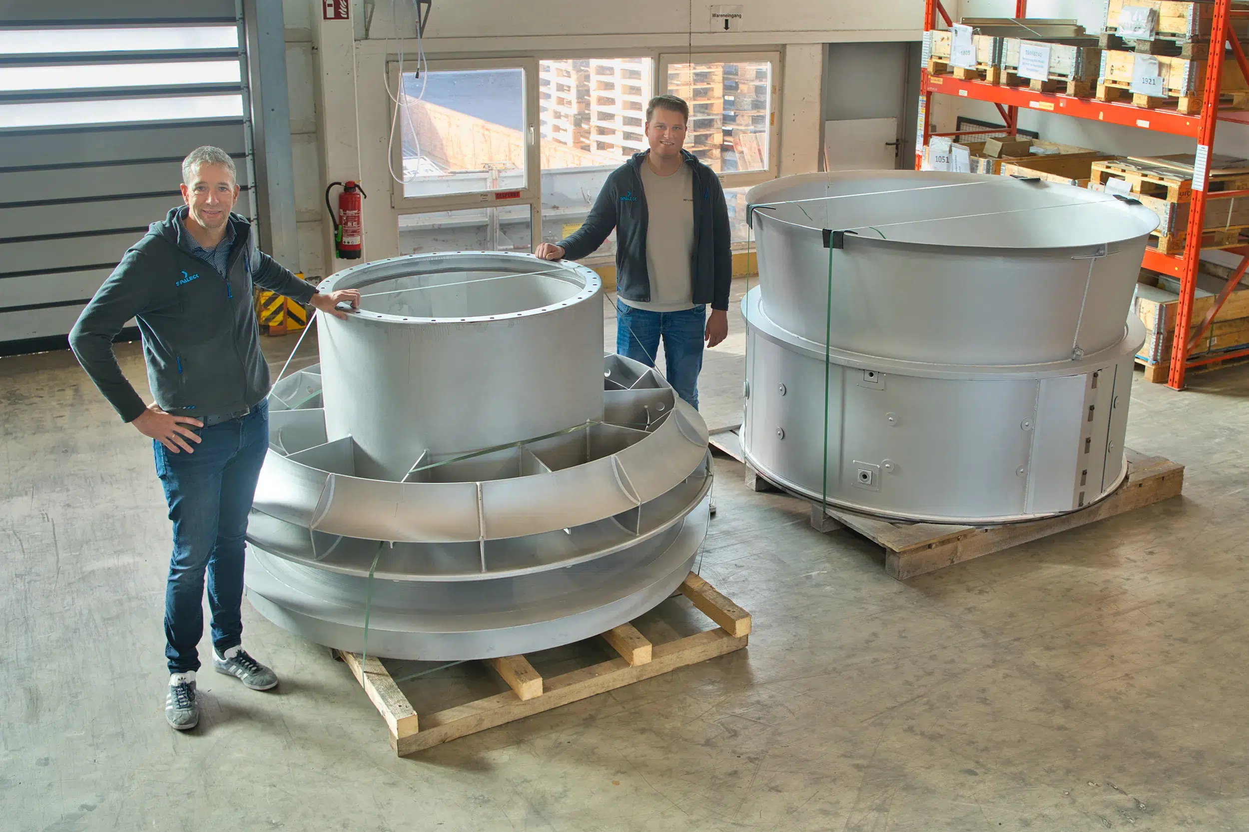 Dois homens estão ao lado de grandes componentes metálicos de uma turbina a gás numa oficina.
