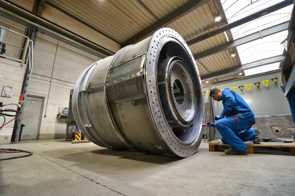 O técnico de construção da SPALECK está ajoelhado junto a um grande conjunto soldado e está a montar peças. O subconjunto faz parte de um grande subconjunto soldado destinado a uma turbina a gás. A imagem mostra a produção na SPALECK.