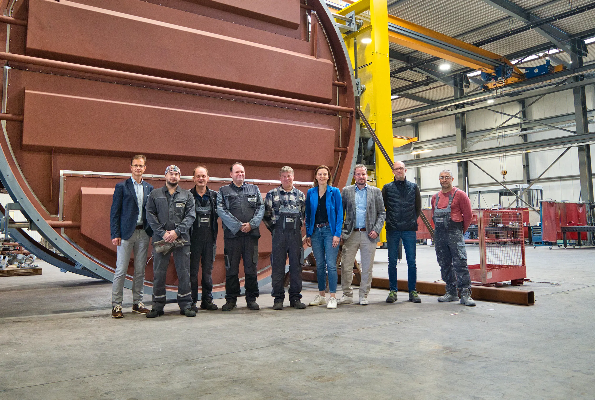 Um grupo de pessoas está em frente a uma grande máquina industrial num pavilhão fabril.